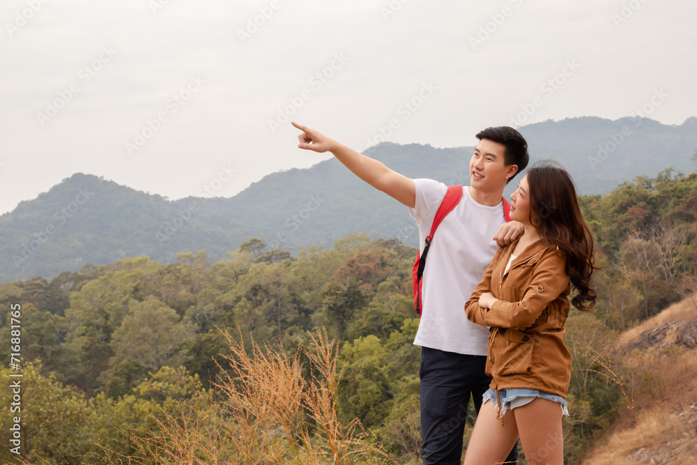 Asian attractive couple travelling together hiking, healthy man and female backpack tourists happy exercise trekking adventure outdoor activity in nature, walking up hill full of golden grass field