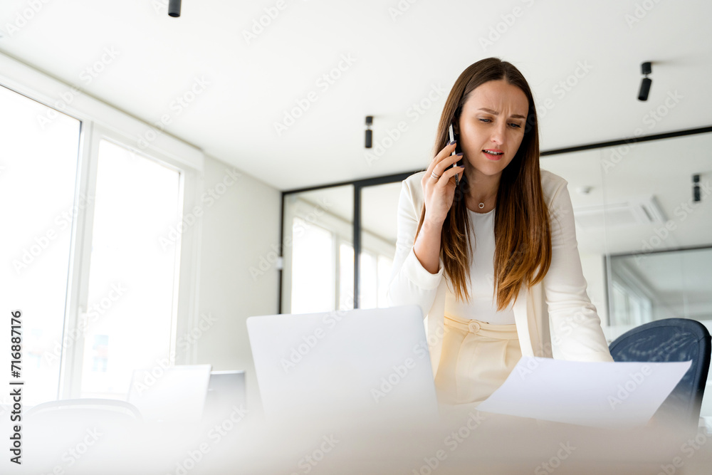 A professional businesswoman speaks on her phone with a concerned expression while examining a document, reflecting the challenging aspects of decision-making and problem-solving in business A professional businesswoman speaks on her phone with a concerned expression while examining a document, reflecting the challenging aspects of decision-making and problem-solving in business