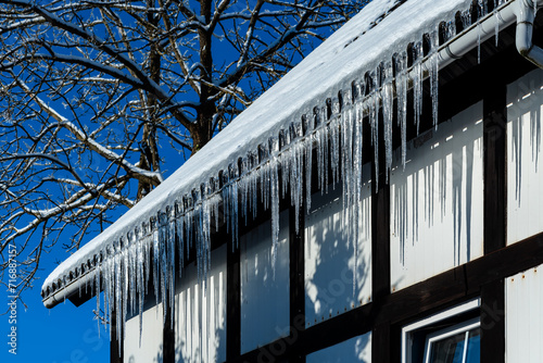 Beautiful icicles hang on the gutter of a historic half-timbered house in Sauerland. Transparent, ephemeral and fragile structures melt and drip in the winter sun. Blue sky and tree in the background. photo