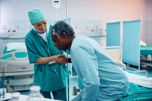 Billede på lærred Black senior patient getting up with help of nurse while recovering after surgery in hospital ward