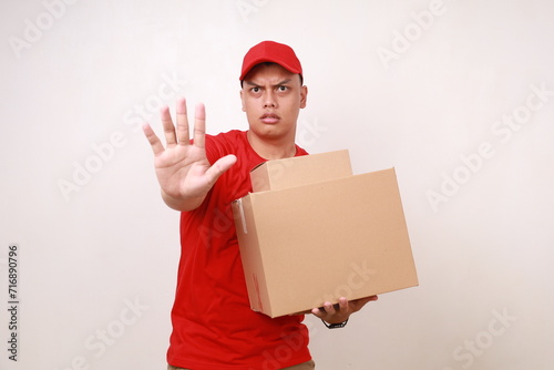 Asian courier man in red holding a cardboard box while showing rejection gesture. Isolated on white photo
