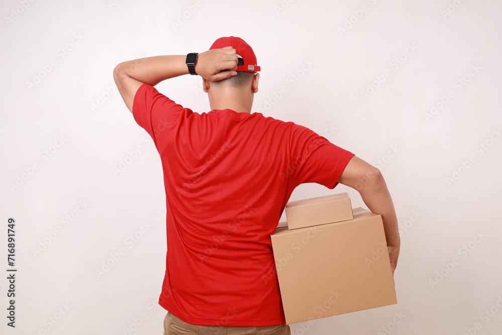 Back view of Confused Asian courier man in red holding head while carrying a cardboard box. Isolated on white Back view of Confused Asian courier man in red holding head while carrying a cardboard box. Isolated on white