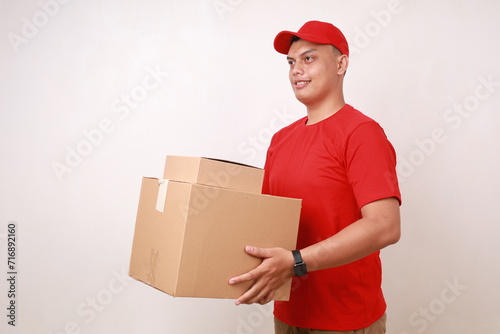 Friendly Asian courier man in red holding and give a cardboard box to sideways. Isolated on white