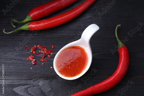 Spicy chili sauce in spoon and peppers on black wooden table, flat lay