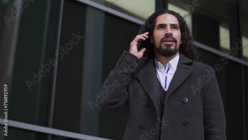 Confident happy mature Latin businessman with long hair looking away while speaking on the mobile phone, outdoors near a modern building photo