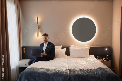 Minimal portrait of elegant businessman using laptop sitting on bed in luxury hotel room with designer interior during work trip, copy space
