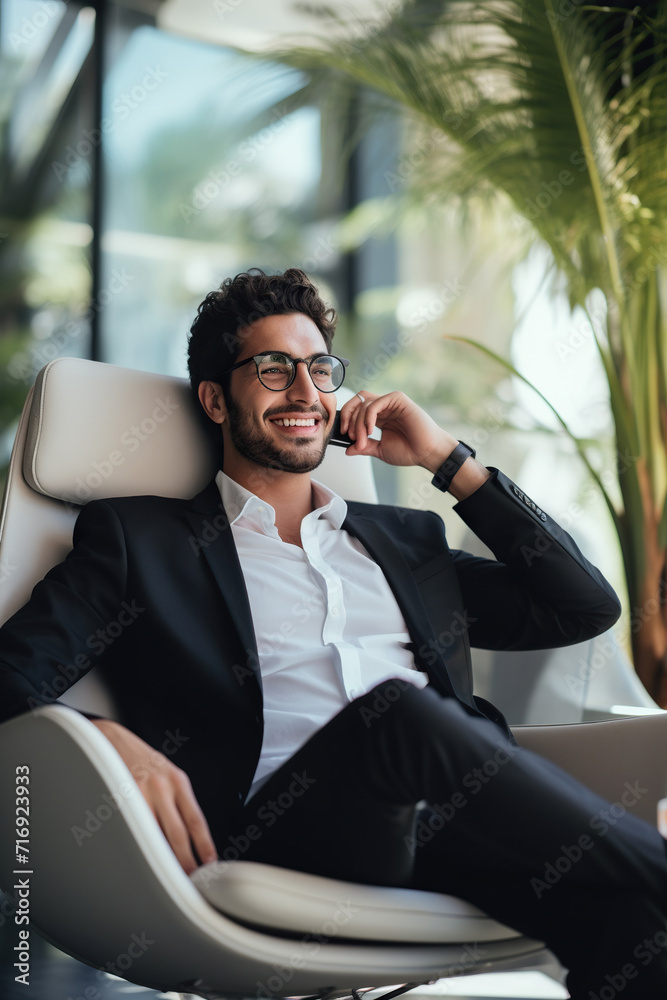 © AI_images - Young Latin businessman wearing eyeglasses and talking over smart phone while relaxing in living room