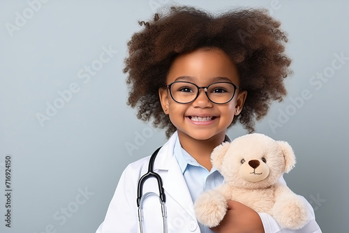 Head shot portrait smiling cute African American girl wearing glasses and white coat uniform with stethoscope pretending doctor looking at camera, playing with fluffy toy patient photo