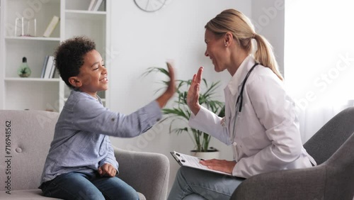 Happy female doctor giving high five to cute little African American boy patient at hospital consultation. A smiling pediatrician will cheer up a small child at the clinic. Healthcare concept.