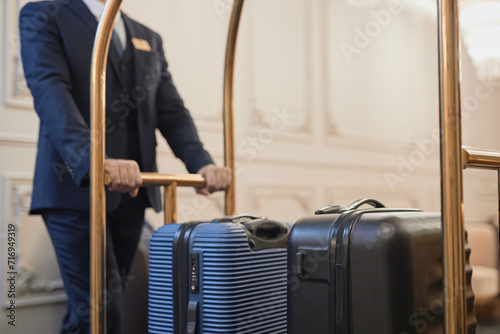 Close up of male porter wearing suit pushing cart with bags and luggage in luxury hotel, copy space photo