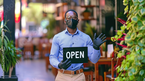 Man with a face mask, wearing a blue shirt, black gloves, and holding a sign that says "OPEN" in front of a restaurant setting with outdoor seating.