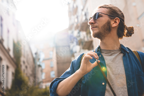 Close up shot of handsome young man in casual clothing looking at view while walking through city streets. Modern caucasian tourist adventurer on a solo trip