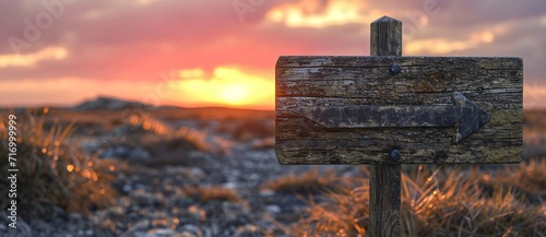 A rustic wooden sign post stands tall against the fiery sky of a winter sunset, marking the way to a tranquil outdoor landscape filled with rolling clouds, golden fields, and vibrant plant life photo
