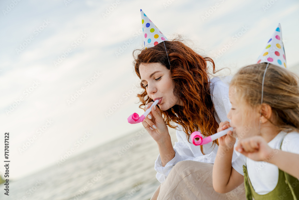 Mother and little girl wearing party hats sitting at beach and blowing whistles.
