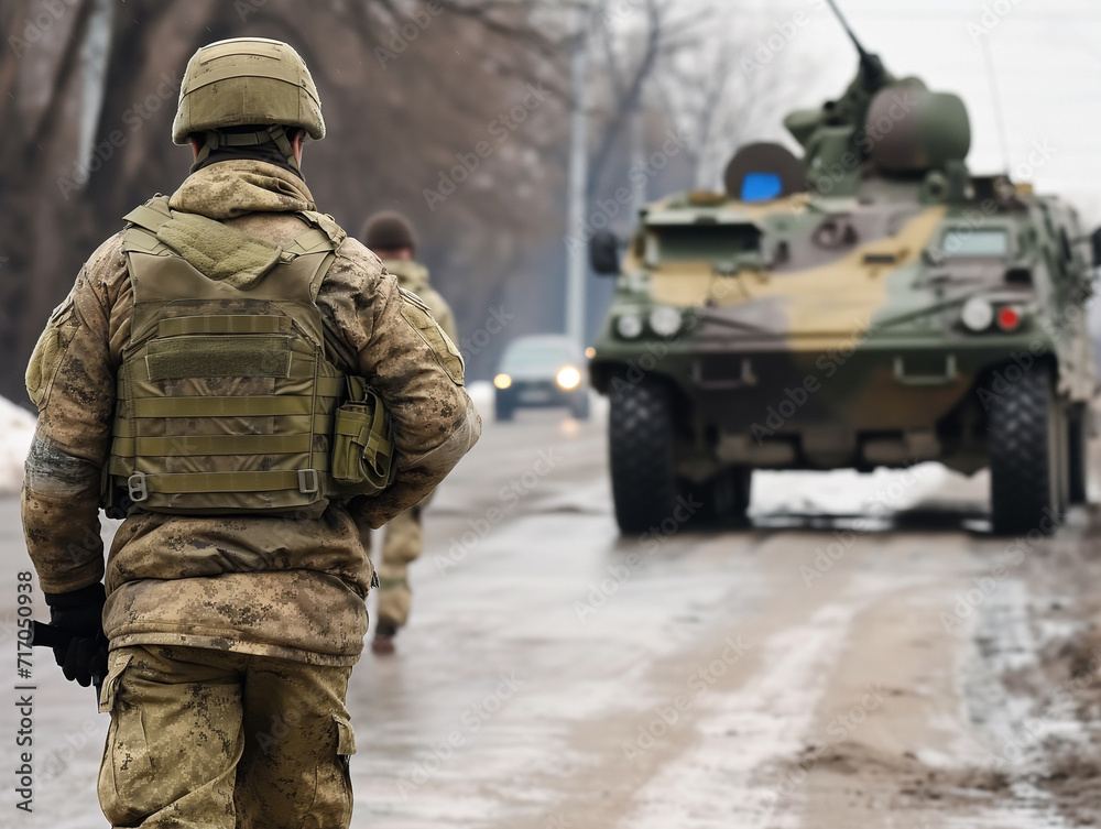 A modern soldier in camouflage with a machine gun stands on a city street against a background of military vehicles. A modern soldier in camouflage with a machine gun stands on a city street against a background of military vehicles.