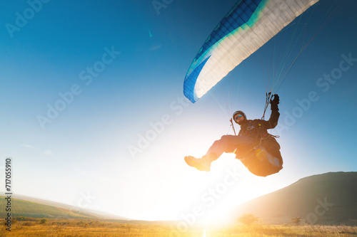 A paraglider takes off from a mountainside with a blue and white canopy and the sun behind. A paraglider is a silhouette. The glider is sharp, with little wing movement.
