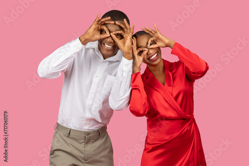 Couple making fun hand gestures over eyes, playful poses, pink background