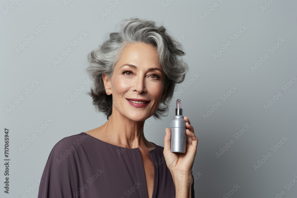 mature woman with silver hair smiles confidently while holding a skincare serum, grey background mature woman with silver hair smiles confidently while holding a skincare serum, grey background