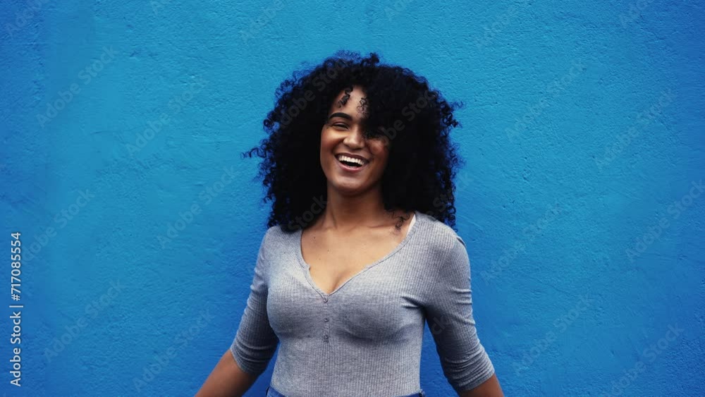 One happy young black Brazilian woman authentic real life laugh and smile standing in outdoor blue backdrop in city street smiling at camera, empowered curly hair African descent person One happy young black Brazilian woman authentic real life laugh and smile standing in outdoor blue backdrop in city street smiling at camera, empowered curly hair African descent person
