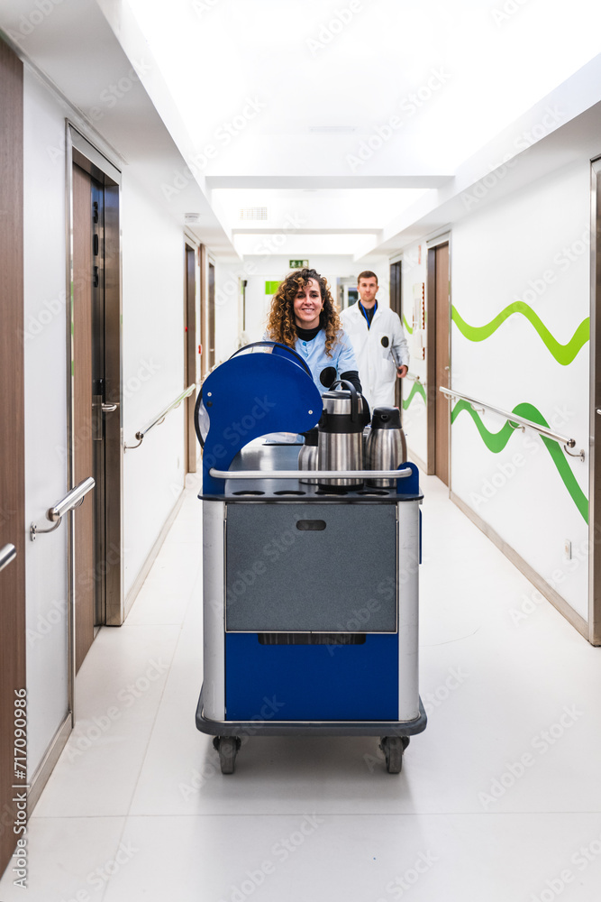 Frontal shot of a cheerful nurse with a coffee service trolley ready to serve patients in a bright hospital hallway.