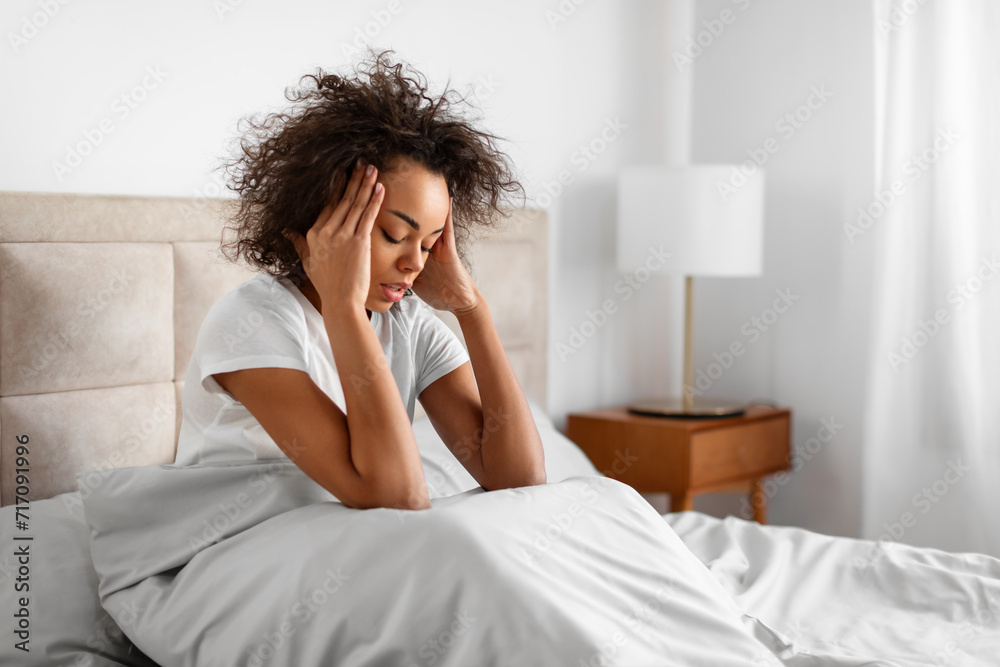 Unhappy black woman sitting in bed touching head having migraine Unhappy black woman sitting in bed touching head having migraine