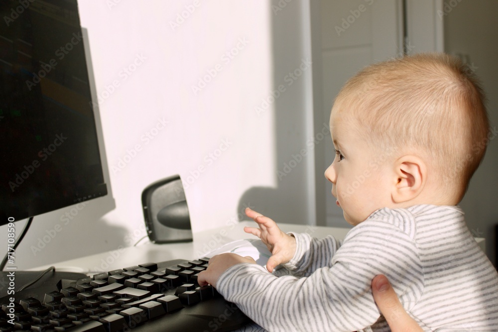 A cute baby boy is sitting at the computer and looks attentively with admiration A cute baby boy is sitting at the computer and looks attentively with admiration