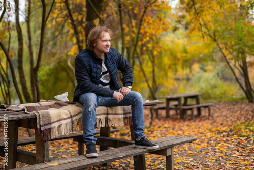 Man Sitting on Bench in Park. A man is seen calmly sitting on a wooden park bench, surrounded by trees and enjoying the tranquility of the environment.