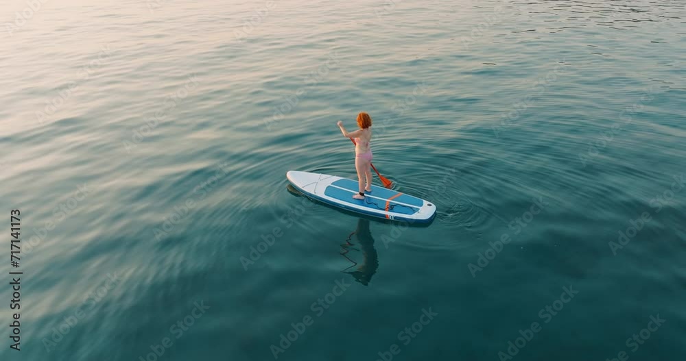 Aerial view young woman floats paddle board on calm blue surface of sea water along coast in Greece at sunset backdrop of hotels in resort town. Summer holiday vacation concept. Lifestyle.Travel
