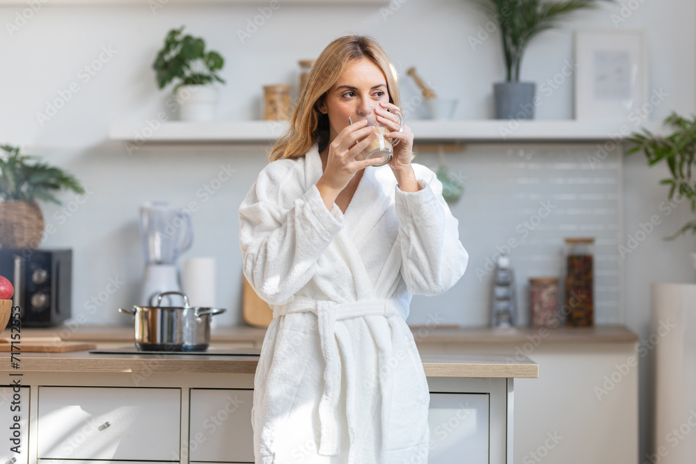 Young woman in bathrobe drinking a cup of coffee while looking forwards in the kitchen Young woman in bathrobe drinking a cup of coffee while looking forwards in the kitchen