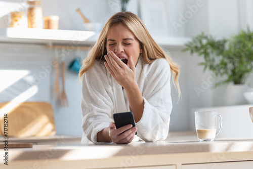 Tired young woman yawning while using her mobile phone in the kitchen at home. photo