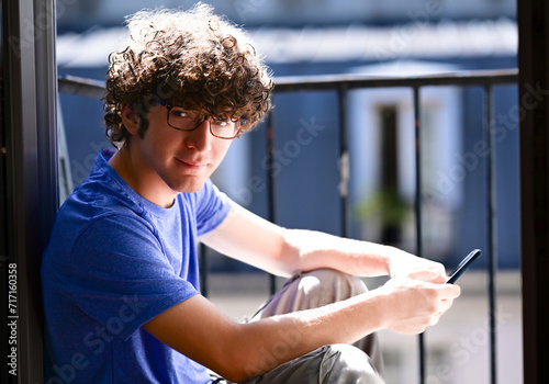 Nice shot of a young Caucasian man with his cell phone. He is sitting on an attic window, he looks into the camera smiling. Natural light illuminates his face and hair. Self-confident and positive. photo