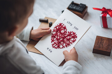 Concept of children craft, handmade gift for Mother's Day. Little boy holding craft card with embroidering in the shape of heart and printed message I love you Mom. Kid's leisure activity, handcraft