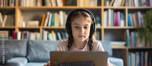 Curious schoolgirl at home, wearing a headset, watches educational videos and answers questions on a laptop. photo