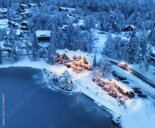 Aerial view of illuminated houses in fairy village in snow, forest, Jasna lake, street lights at winter night. Top view of alpine countryside, snowy pine trees, road at dusk. Kranjska Gora, Slovenia