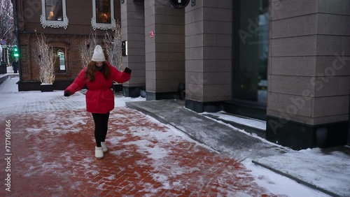 Young woman wearing hat and red trendy winter jacket walking on slippery city sidewalk. Pretty female walking along ice pavement. Danger of season trauma. Shooting in slow motion. photo