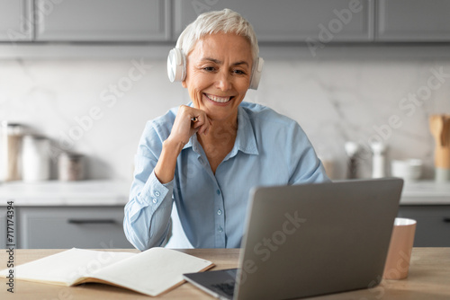 Cheerful senior lady in headphones watching lecture on laptop indoor photo