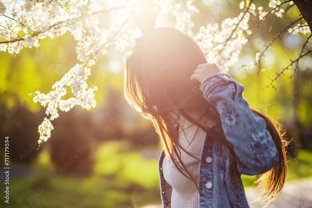 A beautiful young woman with long hair relaxes in a spring park with flowering trees at sunset. - obrazy, fototapety, plakaty 