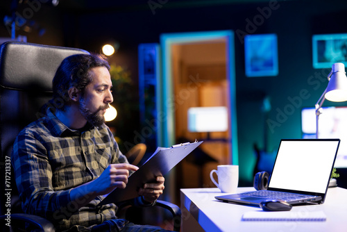 Bearded man is utilizing his digital laptop which is showing a blank chromakey template. Male individual holding clipboard and looking at his device which has an isolated mockup display.