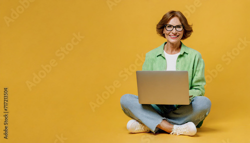 Full body elderly blonde woman 50s years old wear green shirt glasses casual clothes sitting hold use work on laptop pc computer isolated on plain yellow background studio portrait. Lifestyle concept