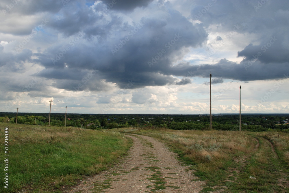 Rural sandy road. The road passes under a gloomy sky with white-gray clouds. Tall green grass grows around it, some of the grass has turned yellow from the sun. A village is visible in the distance.