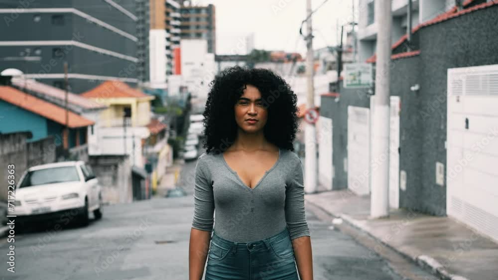 One confident young black woman with curly hair standing outside in urban setting looking at camera with stern solemn expression. South American, 20s person in street portrait face