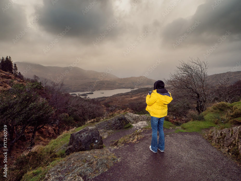 Teenager girl in yellow jacket enjoys stunning nature scene with mountains, river in a valley and dark dramatic sky. Ladies view, Killarney, Ireland, ring of Kerry route. Travel and sightseeing.