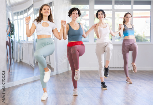 Four happy young women in colorful activewear participating in dance fitness class, lined in row while performing traditional syrtaki moves in bright spacious studio..