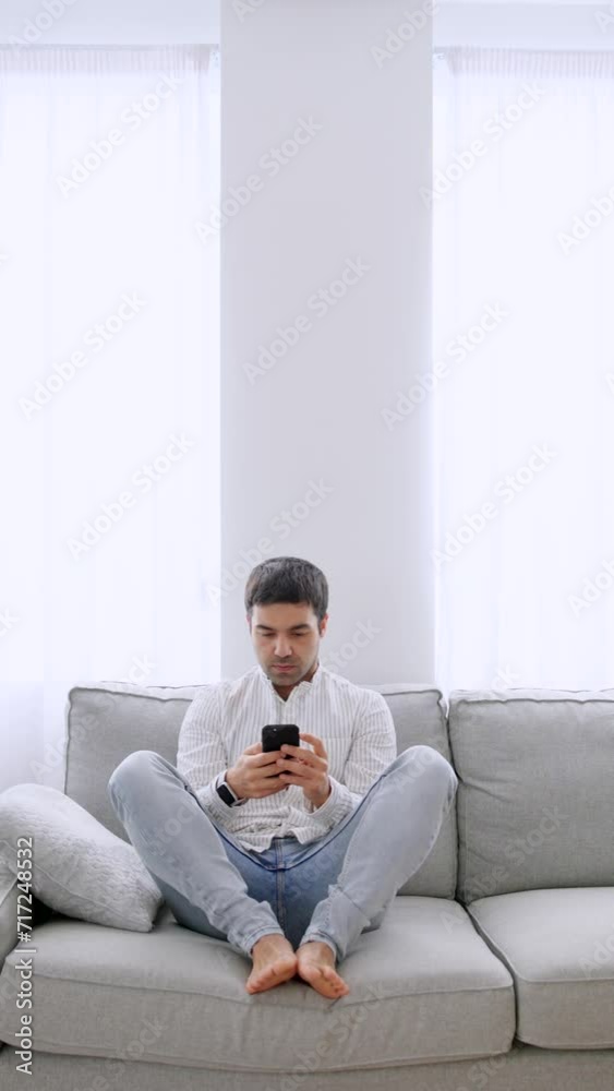 Hispanic young man using phone while sitting on sofa. Person typing on smartphone and chatting on social media Hispanic young man using phone while sitting on sofa. Person typing on smartphone and chatting on social media