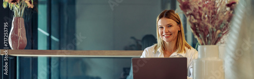 Smiling woman sales manager working on laptop in cozy coworking and looking at camera