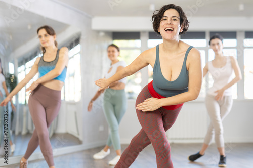 Expressive cheerful young Latina leading upbeat Zumba class for group of women in mirrored dance studio..