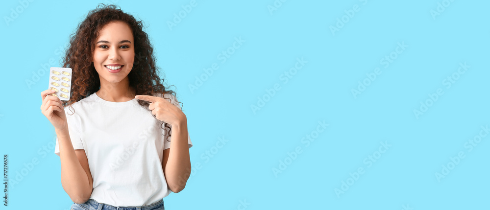 Young African-American holding fish oil pills on light blue background with space for text - obrazy, fototapety, plakaty 
