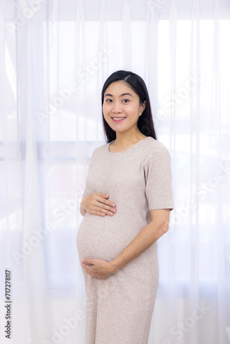 Free Download Portrait of young Asian pregnant woman standing near window with white curtain holding hands on her belly at modern home with copy space. Pregnancy, motherhood, concept.Beautiful pregnancy.Smile. - GFXLoad