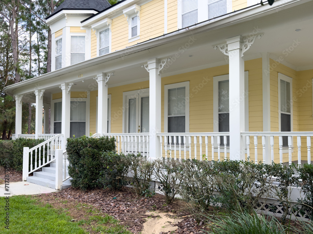 The facade of a large yellow and white colored wooden middle class house with a wraparound porch on the main level. There are multiple double hung glass windows with white curtains and trim in front. 