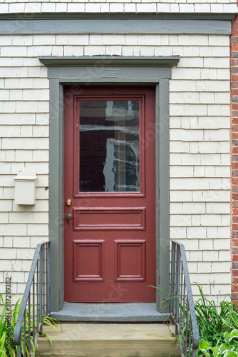 The exterior side entrance to a cream-colored wooden house. There's a closed red door with a half-glass window, wide green trim, metal handrail, steps, and a small letter box affixed to the wall.